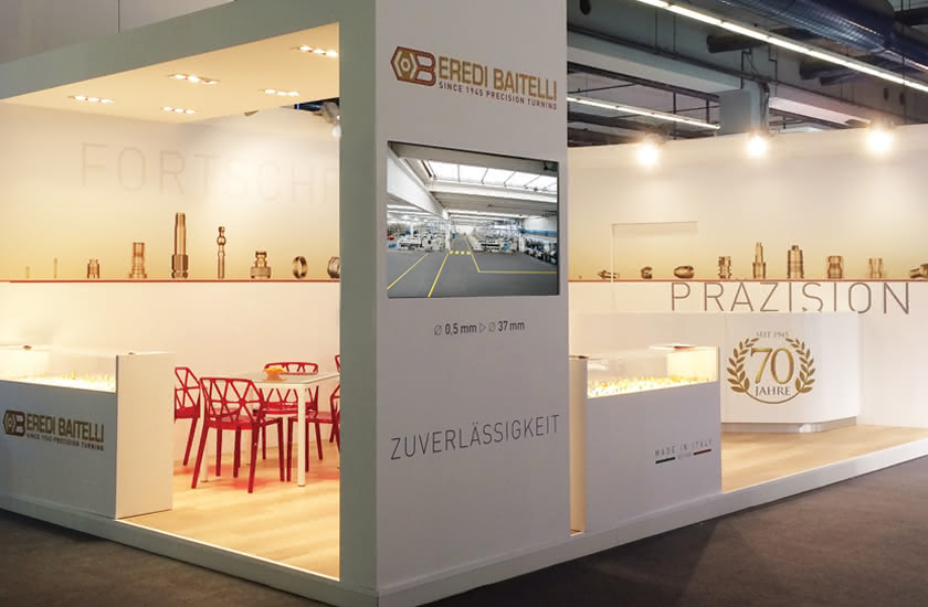 Eredi Baitelli exhibition stand at an international trade fair, with logo, samples of small parts, and multimedia monitor.