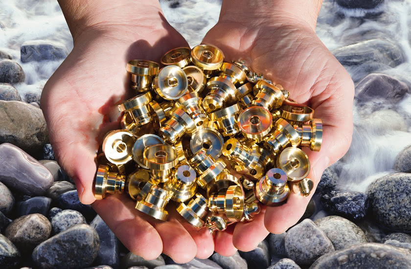Two hands holding a handful of turned brass components on a bed of stones next to a stream.